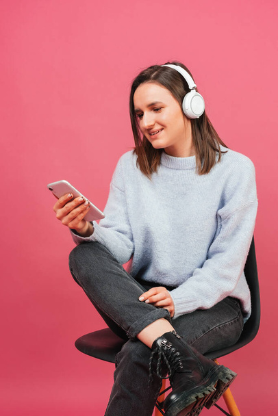 Young woman sits on a chair while browsing music and wearing headphones. Vertical studio shot over pink background. - Fotoğraf, Görsel