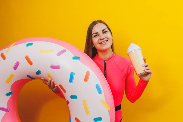 A slim woman in a bright pink swimsuit on a yellow background. She is holding an inflatable pool donut. Beach fashion. - Photo, Image