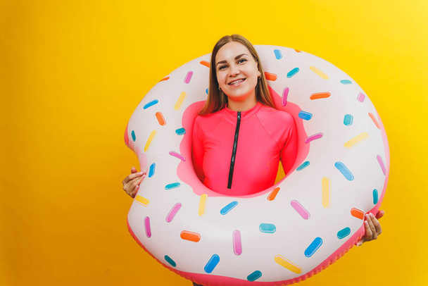 Beautiful young woman in a stylish pink swimsuit with a donut inflatable ring on a yellow background - Photo, Image