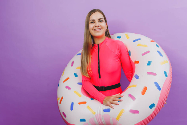Cheerful slender woman with long hair holding an inflatable ring on a purple background. Attractive girl in a pink swimsuit - Photo, Image