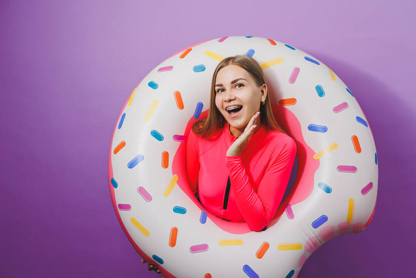 Slim young woman in a stylish pink swimsuit with a donut inflatable ring on a plain background. - Photo, Image