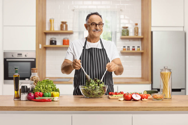 Mature man preparing a salad on a kitchen counter and smiling at camera - 写真・画像