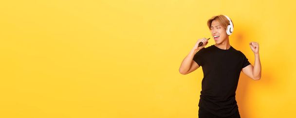 Ξέγνοιαστος όμορφος Ασιάτης με ακουστικά, να παίζει καραόκε, να τραγουδάει σε μικρόφωνο κινητού τηλεφώνου, να στέκεται κίτρινο φόντο. - Φωτογραφία, εικόνα