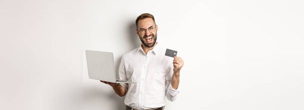 オンラインショッピング。クレジットカードを示すハンサムな男とインターネットで注文するラップトップを使用して、白い背景の上に立って. - 写真・画像