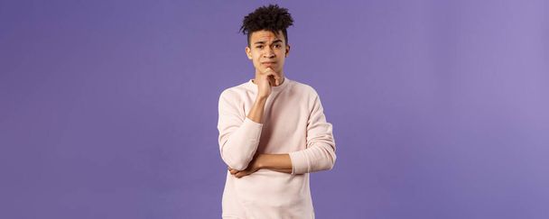 Thoughtful, skeptical young man look judgemental and hesitant at camera while pondering between choices, consider what to do, thinking over purple background, solving troublesome situation. - Photo, Image