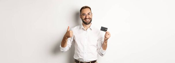 Un bell'uomo che mostra la sua carta di credito e il pollice in su, che raccomanda banca, in piedi su sfondo bianco. Copia spazio - Foto, immagini