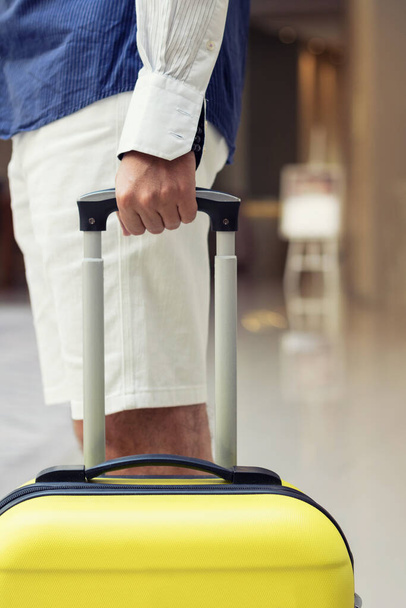 Un hombre con una maleta esperando en el vestíbulo de un hotel. Persona irreconocible con bolsa de viaje de pie en un pasillo. - Foto, imagen