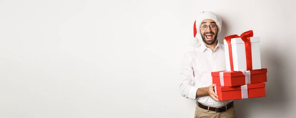 Winterurlaub und Feiern. Glücklicher Kerl bringt Weihnachtsgeschenke, hält Geschenke und trägt Weihnachtsmütze, steht über weißem Hintergrund. - Foto, Bild