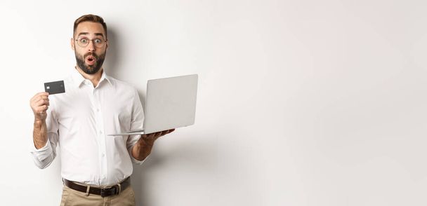 Online shopping. Surprised man holding laptop and credit card, shop internet store, standing over white background. - Photo, Image