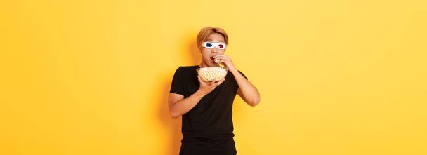 Etkileyici ve heyecanlı Asyalı adam sinemaya gidiyor, 3 boyutlu gözlüklerle film ya da TV dizisi izliyor, patlamış mısır yiyor ve şaşırmış görünüyor, sarı arka planda duruyor.. - Fotoğraf, Görsel
