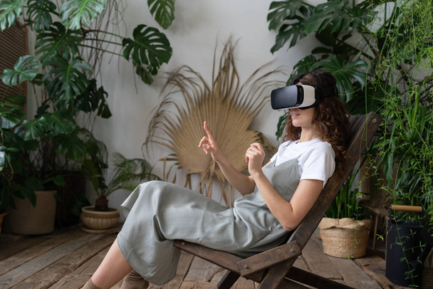 Young woman experiencing virtual reality, sitting among houseplants, curious Hispanic female in augmented reality glasses resting at greenhouse, interacting with AR interface, gardening in cyberspace - Foto, afbeelding