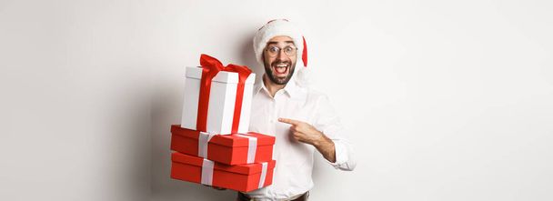 Vrolijk kerstfeest, vakantie concept. Verrast man ontvangt kerstcadeaus, wijzend naar cadeautjes en glimlachend gelukkig, het dragen van santa hoed, witte achtergrond. - Foto, afbeelding