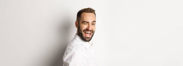 Close-up of cheeky guy with beard, turn face at camera and winking with smile, standing over white background. - Fotó, kép