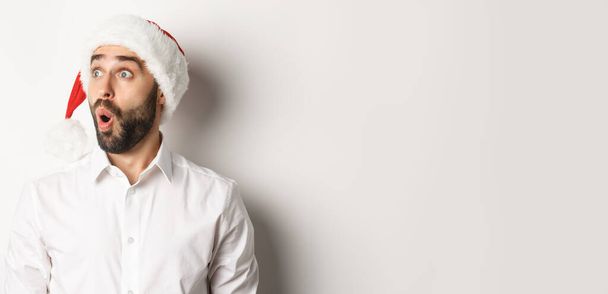 Κοντινό πλάνο του καταπλήσσει γενειοφόρος άνθρωπος στο καπέλο santa, κοιτάζοντας αριστερά με έκπληξη πρόσωπο, τα Χριστούγεννα προσφοράς promo, στέκεται πάνω από το λευκό φόντο. - Φωτογραφία, εικόνα