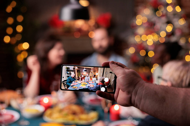 Family christmas celebration photo on smartphone screen, friends eating at festive dinner table on blurred background. Xmas holidays celebrating, hand holding mobile phone close up - 写真・画像