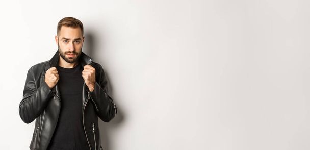 Image of handsome and confident man putting on leather biker jacket, standing against white background. - Foto, Imagen
