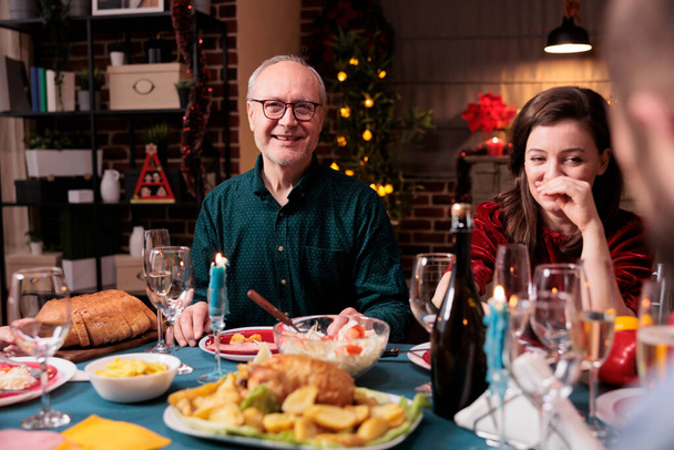 Szczęśliwa kobieta świętująca święta z rodziną, śmiejąca się przy świątecznym stole z tatą na przyjęciu domowym. Ojciec i córka jedzą razem naczynia świąteczne, uśmiechają się, bawią, patrzą w kamerę - Zdjęcie, obraz