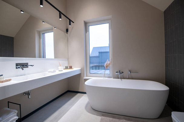 Ceramic bathtub installed by a large window before the wall-mounted mirror with the lighting fixtures and the drop-in sink - Foto, Imagen