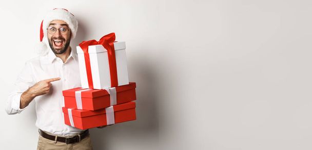 Счастливого Рождества, праздничная концепция. Удивленный человек получает рождественские подарки, указывая на подарки и улыбаясь счастливый, в шляпе Санты, белый фон. - Фото, изображение