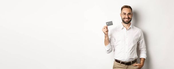 Przystojny biznesmen pokazujący swoją kartę kredytową, wyglądający na zadowolonego, stojący na białym tle. - Zdjęcie, obraz