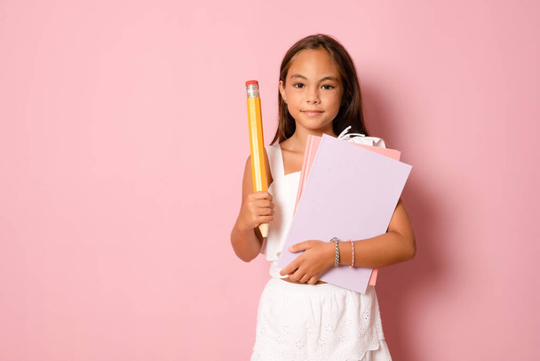 Cute pretty child girl with books on pink background holding a big pencil. - 写真・画像