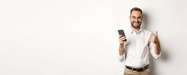 Ικανοποιημένος επιχειρηματίας δείχνει αντίχειρες επάνω μετά τη χρήση του κινητού τηλεφώνου, στέκεται ευχαριστημένος πάνω από λευκό φόντο. - Φωτογραφία, εικόνα