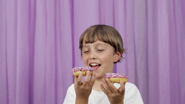 Jongen van 9 jaar oud bijt een donut op een paarse achtergrond. Hoge kwaliteit 4k beeldmateriaal - Video