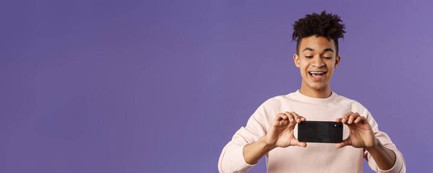 Portrait of amazed, excited young man seeing something interesting, stream concert to his internet social network profile, taking photo or recording video with mobile phone, purple background. - Photo, Image