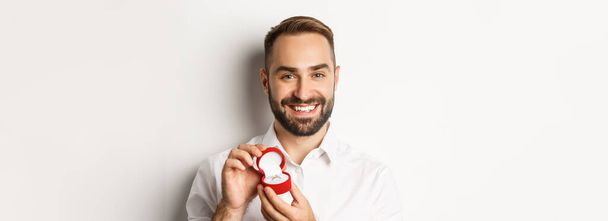 Közelkép egy boldog jóképű férfiról, aki lánykérést tesz, jegygyűrűt tart a dobozban és mosolyog, megkéri a kezét, fehér háttér.. - Fotó, kép