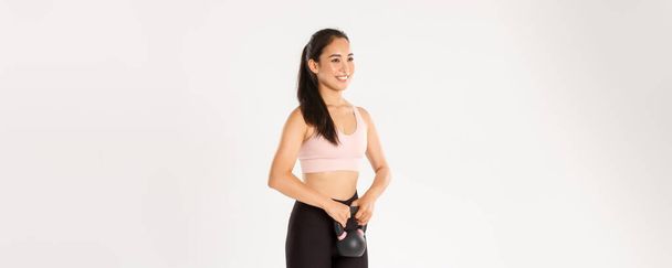 Sport, wellbeing and active lifestyle concept. Smiling strong and slim asian fitness coach, workout trainer or girl in gym lifting kettlebell, do squats with weight, standing white background. - Zdjęcie, obraz