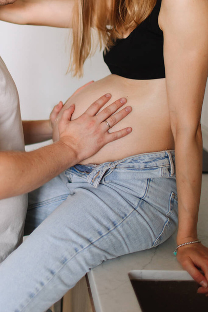 Una chica embarazada con el pelo rubio en jeans desabotonados se sienta a la mesa, y su marido toca su vientre embarazada. - Foto, imagen