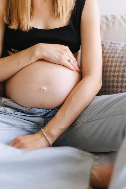 A pregnant girl with blond hair in unbuttoned jeans hugs her pregnant belly. - Zdjęcie, obraz