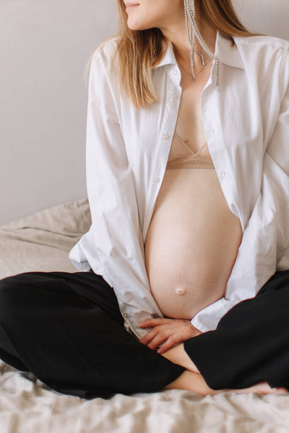 Une fille enceinte portant une chemise blanche et un pantalon est assise avec un ventre nu et enceinte. Cheveux blonds, boucles d'oreilles volumineuses. - Photo, image