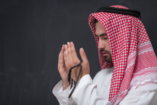Młody arabczyk w tradycyjnym ubraniu modlący się tradycyjnie do Boga trzyma ręce w modlitewnym geście przed czarnym tłem. Wysokiej jakości zdjęcie - Zdjęcie, obraz