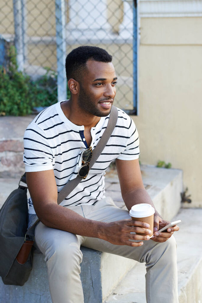 Profitant d'un café à emporter rapide. Un bel homme d'affaires afro-américain vêtu de façon décontractée assis dans la ville et utilisant son téléphone portable tout en buvant du café - Photo, image