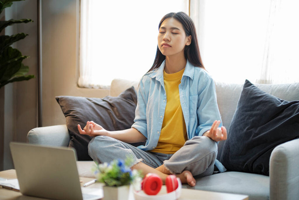 Klidná žena v neformálním vzhledu medituje, cvičí jógu, dýchá a soustředí se na gauč, aby snížila stres, odpočívá po práci doma. Vědomí a koncept mírové meditace. - Fotografie, Obrázek