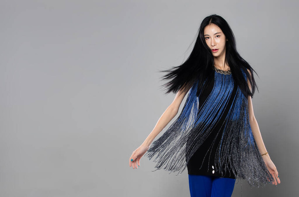 Ázsiai transznemű nő hosszú fekete egyenes hajjal, szél fúj dobja a levegőt. Női spin viszont, és viseljen kék divat érzéki szexi ruha, szürke háttér elszigetelt másolás tér - Fotó, kép