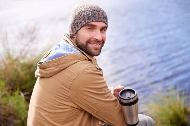 Its great out here. Portrait of a handsome man sitting by a lake holding a thermos - Photo, Image