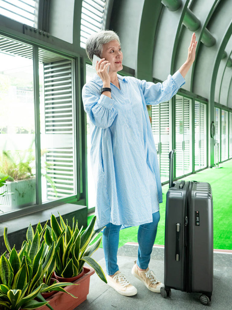 Portrait of senior adult elderly asian woman 60s smiling standing with suitcase luggage bag and using smartphone for travel concept. - Fotoğraf, Görsel