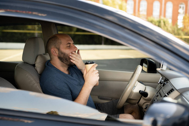 Κουρασμένος άνδρας χασμουριέται στο μπροστινό κάθισμα του αυτοκινήτου κρατώντας καφέ για να πάει. Μεταφορά έννοια του ατυχήματος στέρηση ύπνου - Φωτογραφία, εικόνα