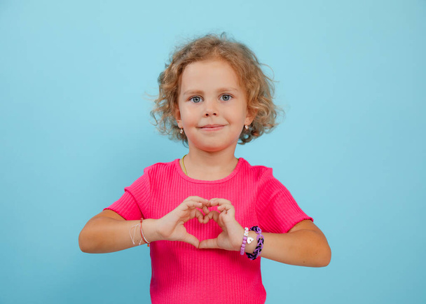 Small little expressive, confident, playful, positive curly blonde girl in pink shirt showing heart sign by hands to camera on blue studio background. Love, care and family protection. Copy space - 写真・画像