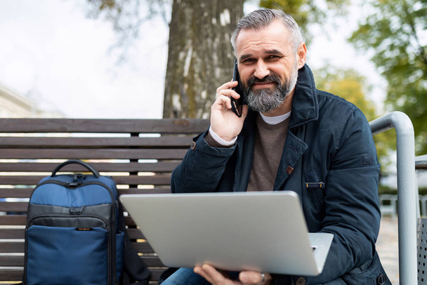 egy idős üzletember mobiltelefonon beszél, miközben egy laptopon dolgozik online egy parkban egy padon.. - Fotó, kép