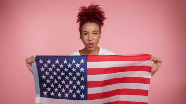Sorridente afro-americano patriota rossa donna riccia in possesso di bandiera americana dice USA guarda telecamera. Ritratto femminile in piedi con bandiera statunitense su sfondo rosa isolato. 4 luglio Giorno dell'indipendenza americana - Filmati, video