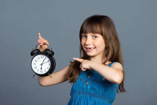 A little preschooler girl smiles and holds a round alarm clock in her hands and shows the time with her index finger. Isolated on a gray background. Time to study and don't be late for school. High quality photo - Photo, image