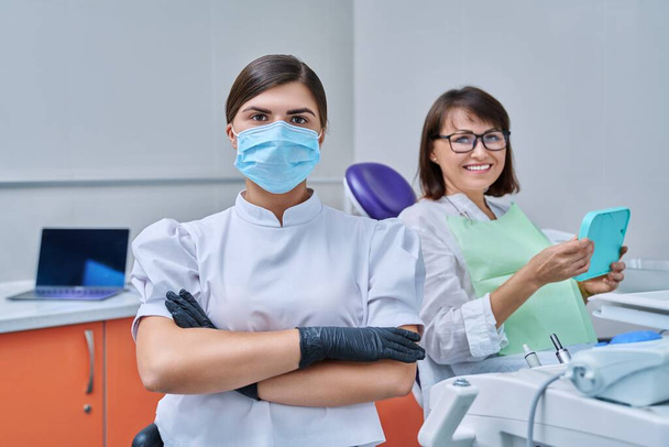 Portrait of young female doctor dentist looking at camera with mature woman patient sitting in dental chair. Dentistry, hygiene, treatment, medicine, dental health care concept - Foto, Imagem