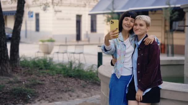 Лесбийская пара делает селфи, используя телефон, делая селфи, стоя в центре города, чувствуя себя счастливыми вместе. Молодая счастливая лесбийская пара делает фото на улице - Кадры, видео