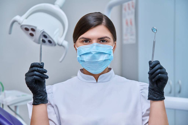 Headshot portrait of female dentist wearing facial mask with instruments for examining teeth. Doctor looking at camera, posing in dental office. Dentistry, medicine, treatment, dental health care - Foto, Imagem