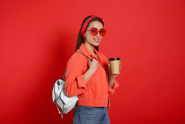 Beautiful young woman with stylish leather backpack and cup of coffee on red background - Fotó, kép