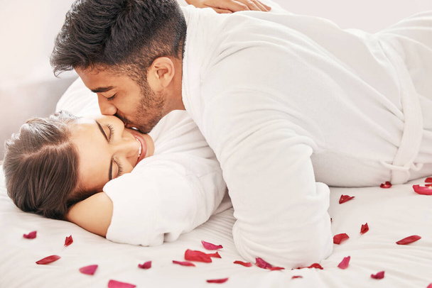Hotel, happy and love couple on a holiday for a romantic anniversary trip with happiness. Kiss, hug and passion of young lovers smile on a bed of rose petals in a bedroom together with happiness. - Photo, Image
