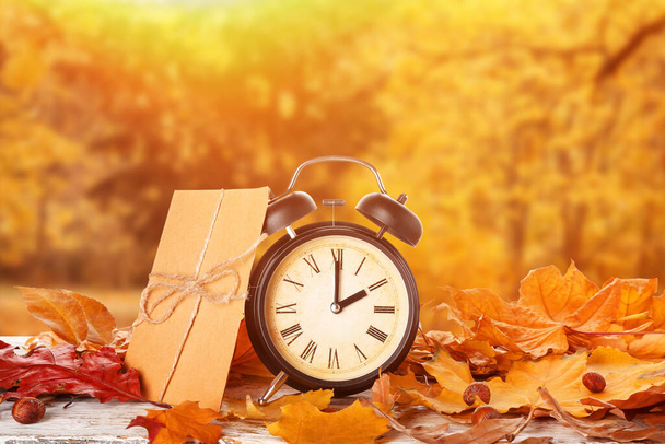 Alarm clock, envelope and autumn leaves on table in park. Daylight saving time end - Photo, Image